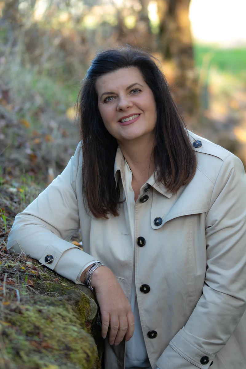 Miriam König im hellen Mantel, an eine moosbewachsene Mauer gelehnt.